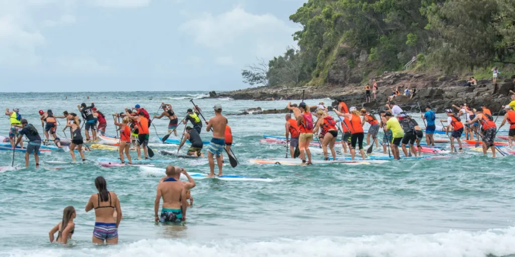 A large group of people paddleboarding in the ocean near a rocky, tree-covered shoreline, with some swimmers and children playing in the shallow water in the foreground.