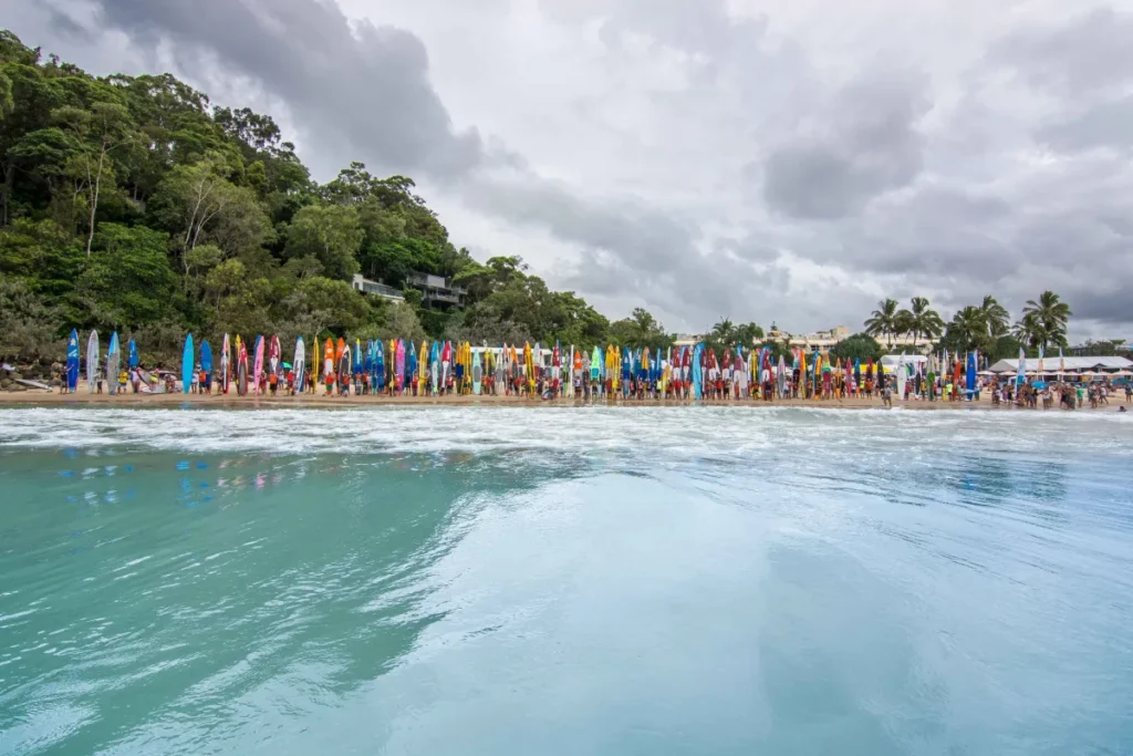 A row of colorful surfboards stands upright in the sand along a crowded beach, with people gathering near the shore and lush green trees in the background under a cloudy sky.