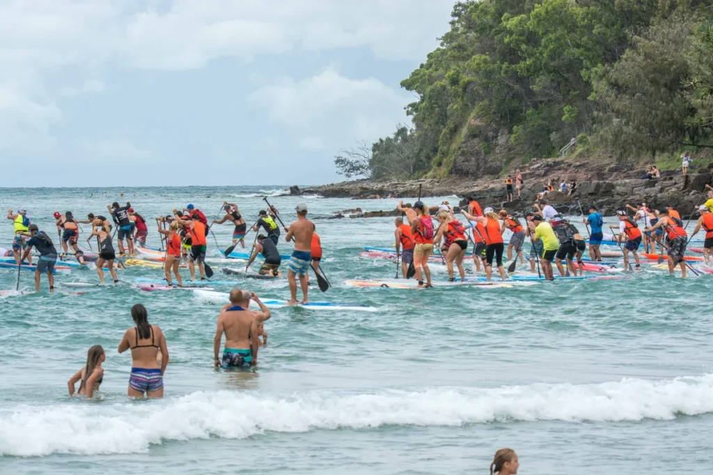 A large group of people in orange vests paddleboard together near a rocky shoreline, while others swim and stand in the shallow water on a cloudy day. Lush green trees are visible along the coast.