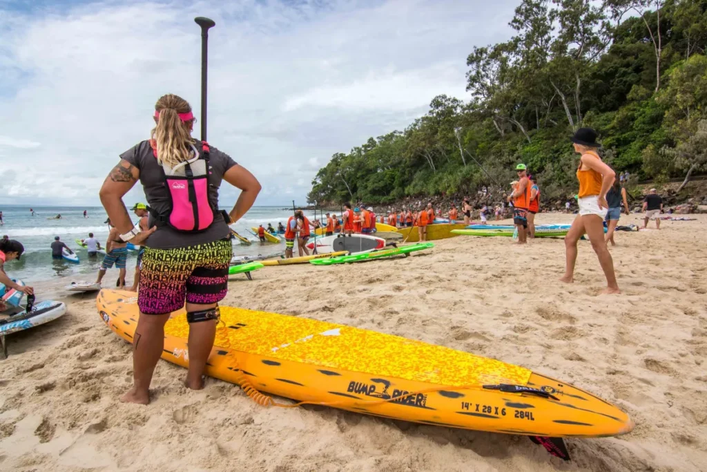 People gather on a sandy beach with paddleboards. One person in colorful shorts and a pink vest stands in the foreground, while others, many in orange vests, prepare near the water. Trees line the background.