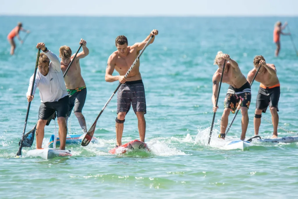 Five men are stand-up paddleboarding in the ocean, competing in a race. They are paddling vigorously, wearing swim trunks, and the water splashes around their boards. In the background, two more paddlers are visible.