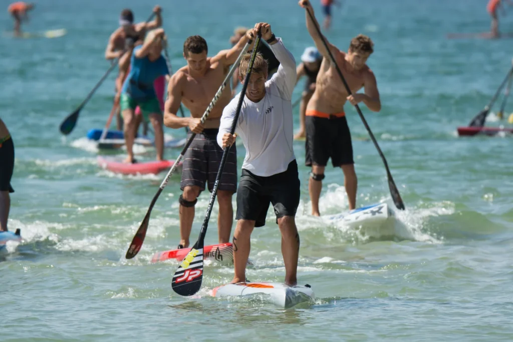 A group of people paddleboarding in the ocean, racing forward while standing upright on their boards and using paddles, with one person leading in the foreground.