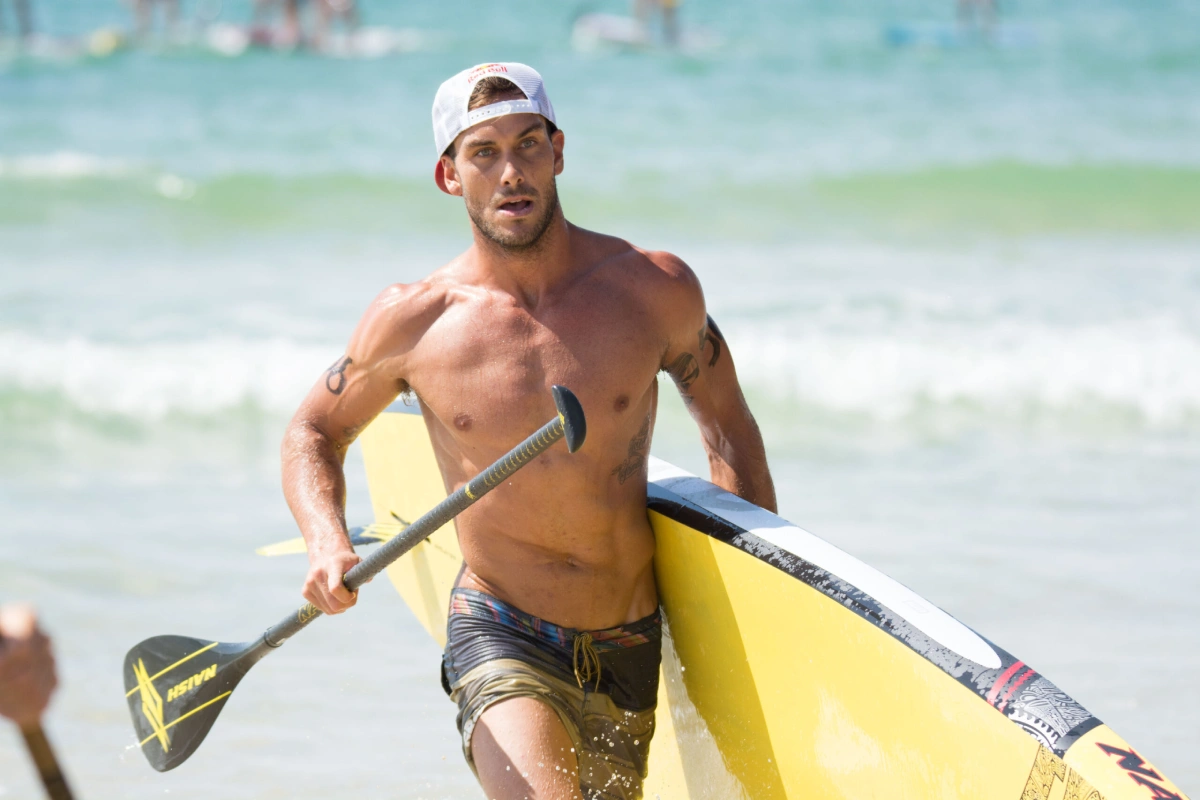 A muscular man wearing a backward white cap carries a yellow paddleboard and a paddle while running out of the ocean onto the beach, with people and water in the background.