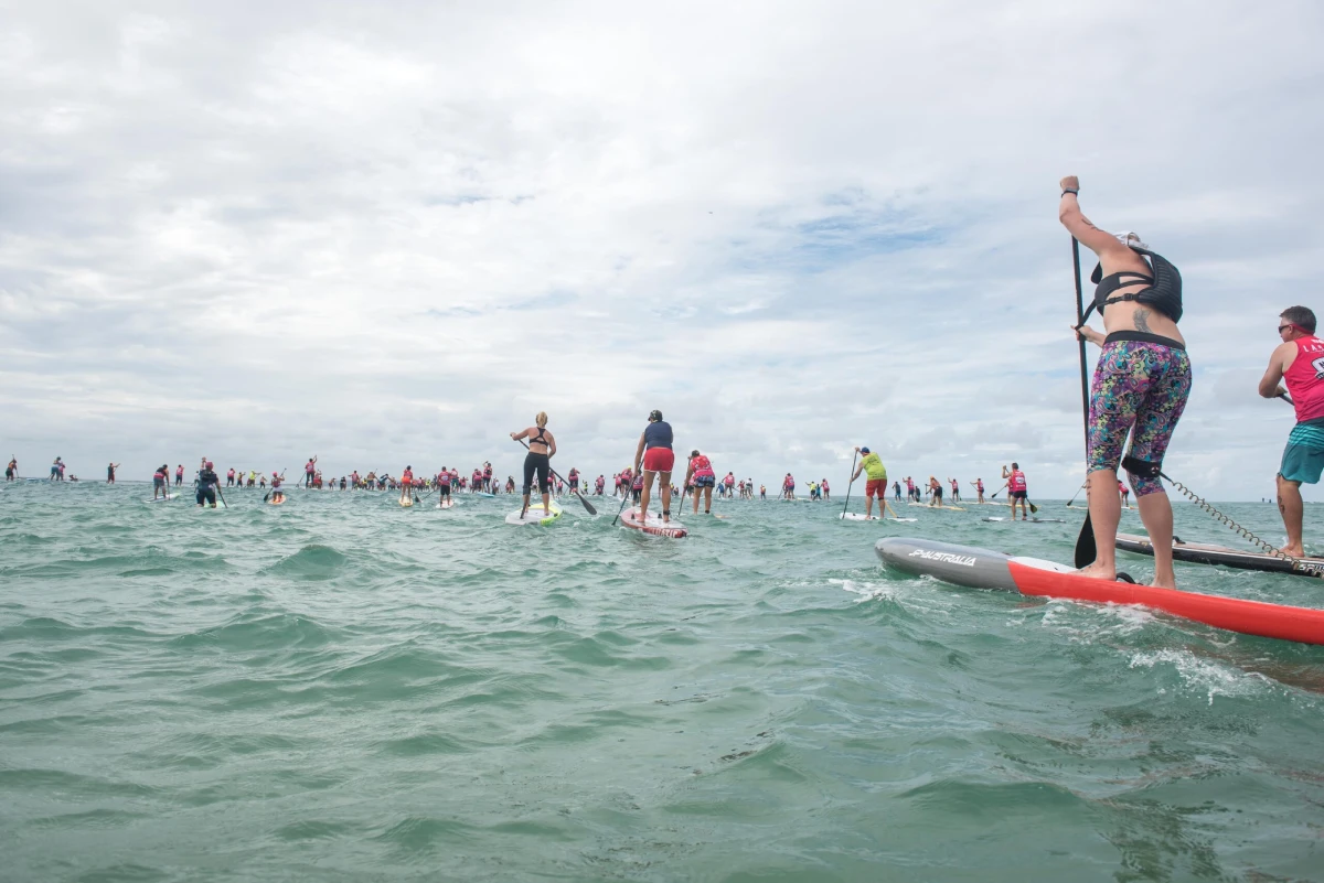 A large group of people paddleboarding on the ocean under a cloudy sky, with some close to the camera and many more in the distance.