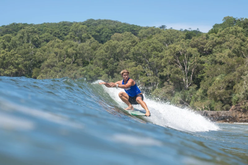 A person in a blue outfit is surfing on a wave near a lush, green forested coastline under a clear blue sky.