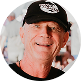Smiling older man wearing a black "World Surf" cap and black shirt, standing in a brightly lit, blurred outdoor setting.