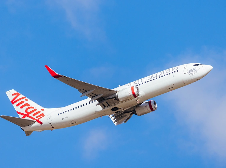 A Virgin Australia airplane with red accents is flying against a bright blue sky. The plane is seen from below, with its landing gear retracted and the airline's logo visible on the tail and fuselage.
