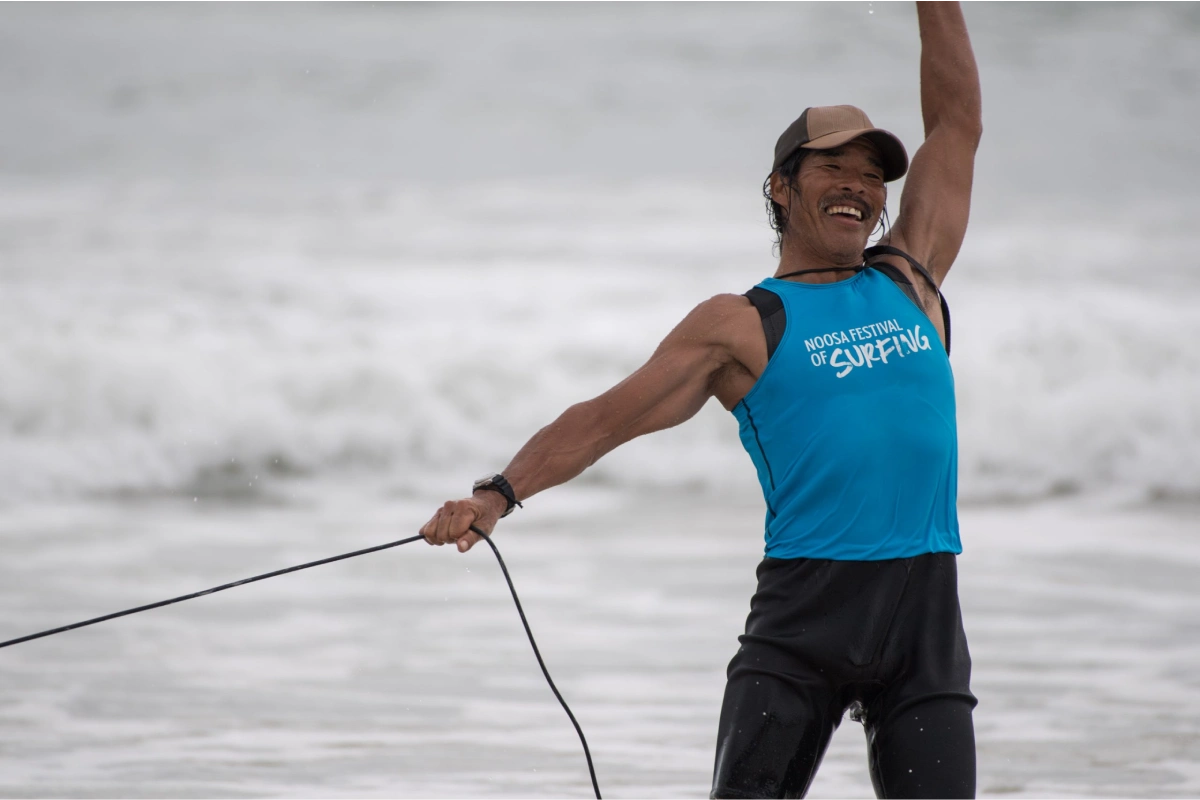 A man wearing a blue "Noosa Festival of Surfing" tank top and black hat stands on the beach with one arm raised, smiling, and holding a surfboard leash. Waves and cloudy skies are in the background.