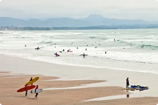 Surfers carrying boards on a sandy beach, with many people surfing and waiting in the waves. The sky is overcast, and mountains are visible in the background.