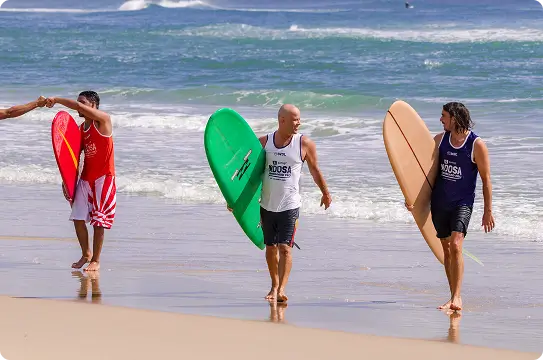 Three men in tank tops and swim trunks walk along the beach carrying surfboards. Two shake hands while waves roll in behind them under a clear blue sky.