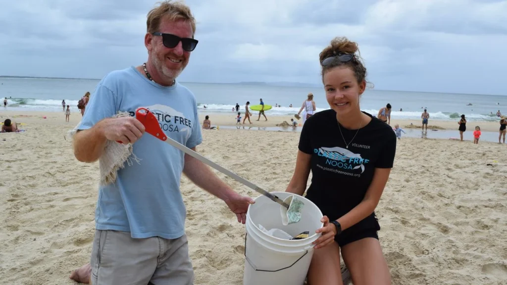 Two people on a sandy beach collect trash in a white bucket, smiling at the camera. The man uses a grabber tool, and the woman holds the bucket. Other beachgoers and the ocean are visible in the background.