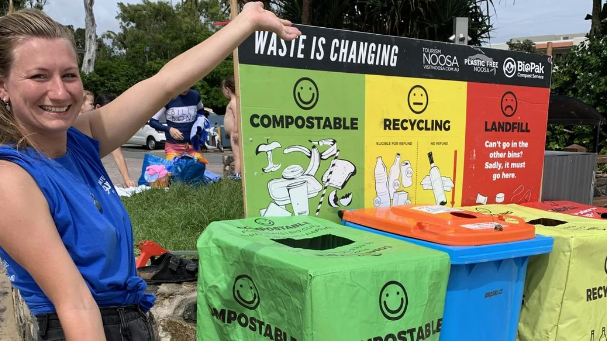 A smiling woman stands beside a waste sorting station with labeled bins for compostable, recycling, and landfill items under a “Waste is Changing” sign at an outdoor event.