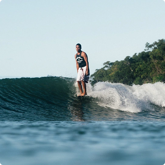 A man in a black tank top and white shorts is surfing on a small wave near a tree-covered shoreline under a clear sky.
