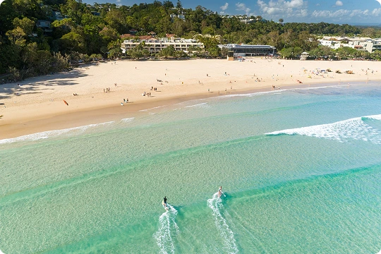 Aerial view of a sandy beach with gentle waves, two people paddleboarding in clear water, and buildings surrounded by trees in the background.