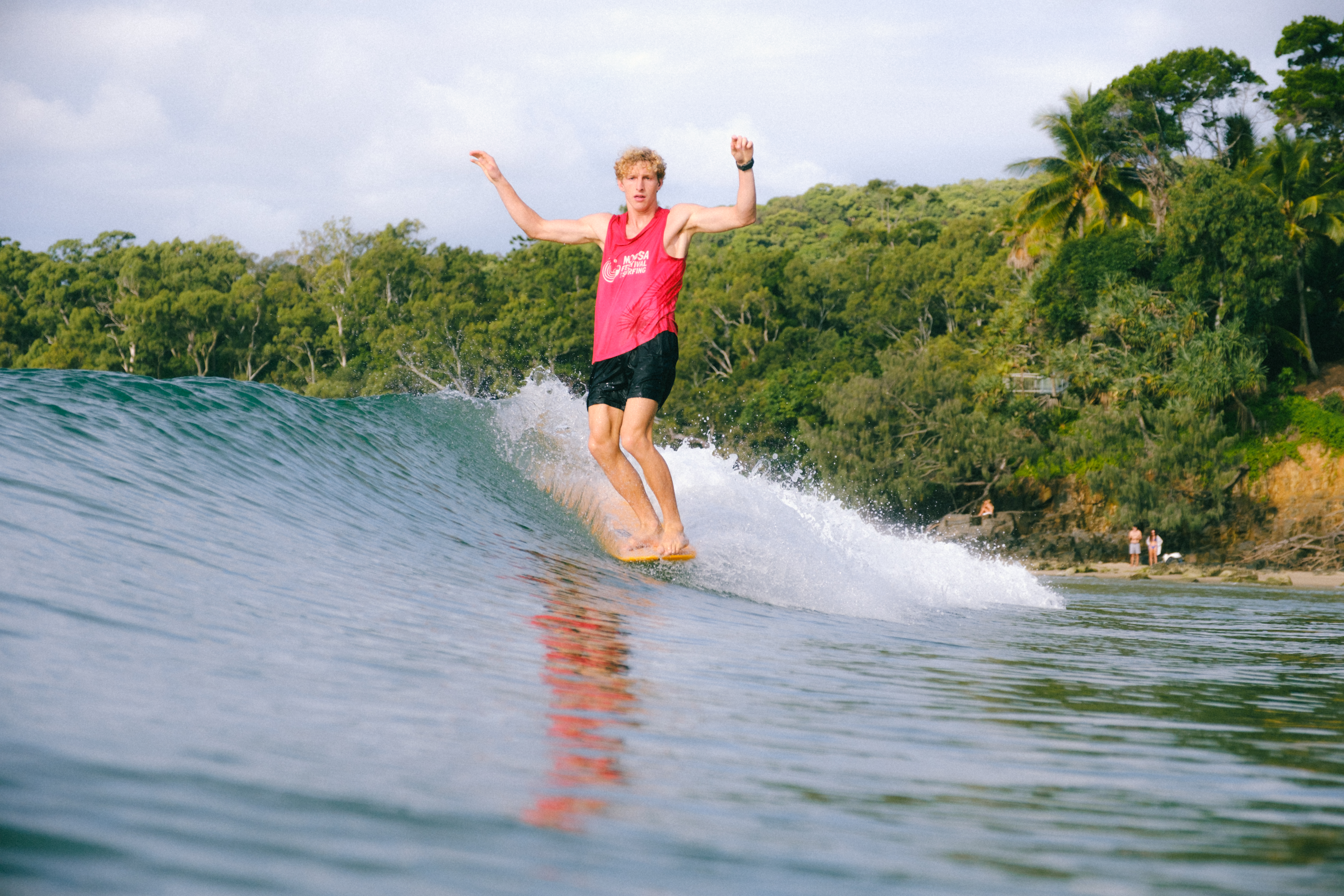 A surfer in a pink tank top rides a wave near a forested shoreline.